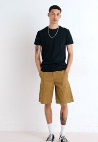 Tricou negru cu guler rotund, pantaloni scurți cargo bej cu buzunare laterale, șosete albe și adidași negri. Modelul stă în fața unui fond alb.