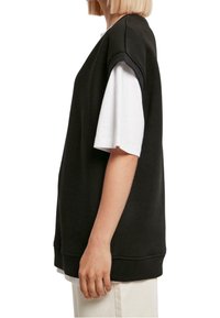 Czarna, oversize bluza z opuszczonymi ramionami, teksturowana tkanina i ściągacz na dole. Nosi się ją na białą koszulę, co tworzy kontrastujące kolory.