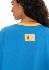 Žena s tmavými kudrnatými vlasy má na sobě modrou košili se žlutým límečkem a nášivkou na zadní straně, která zobrazuje citrón a text "Club Tropicana."