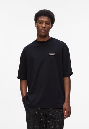 T-shirt nera oversize in cotone, con collo rotondo e maniche corte. Include una piccola etichetta con il logo sul davanti.