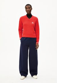 Vrouw in een rode kabelgebreide trui over een zwarte coltrui, met een marineblauwe wijde broek en witte schoenen, staand met haar handen in haar zakken.