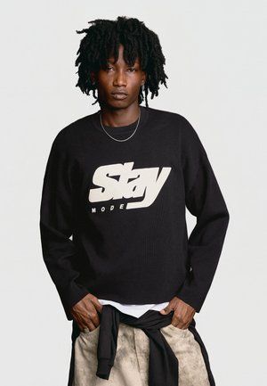 Jeune homme avec des dreadlocks portant un pull noir avec le texte « "Stay Mode" », un pantalon beige et une veste noire nouée autour de la taille.