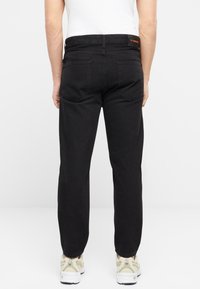 Los jeans de mezclilla negros presentan un diseño de pierna recta con dos bolsillos traseros y una etiqueta que dice "ZT Premium". Se usan con zapatillas beige.