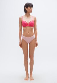 Γυναίκα που στέκεται ξυπόλυτη, φοράει κόκκινο δαντελωτό σουτιέν και ανοιχτό ροζ εσώρουχα, κοιτάζοντας προς τα αριστερά της, μπροστά σε απλό λευκό φόντο.