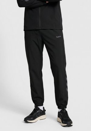 Person iført sort lynlåstrøje og sorte Hummel træningsbukser med elastiske manchetter og sorte sneakers med hvide såler.