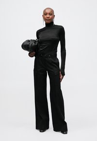 Schwarzes Langarmoberteil, kombiniert mit hoch taillierten, weiten schwarzen Hosen. Das Outfit wird von einer strukturierten Tasche und auffälligen Gürteldetails ergänzt.
