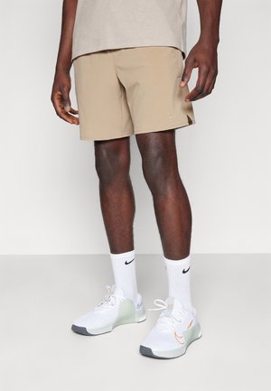 Homme portant un short beige Nike, des chaussettes blanches Nike et des chaussures de sport blanches Nike, debout devant un fond clair uni.