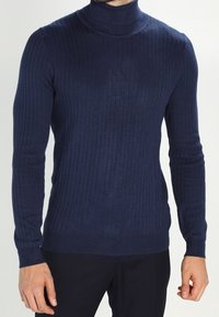 Man i en åtsittande marinblå ribbad polotröja med långa ärmar och mörka byxor, stående mot en enfärgad bakgrund.