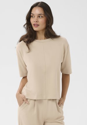 Femme aux cheveux longs et ondulés portant un haut beige à manches courtes et un pantalon assorti, les mains dans les poches, regardant vers la gauche sur un fond uni.