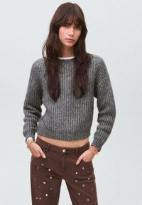 Sivi pleteni pulover s rebrastom teksturom i skraćenim dizajnom, uparen s smeškim hlačama koje imaju raspršene metalne točke.