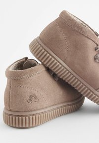 Bottes chelsea en daim marron clair avec doublure douce, bout rond, semelle en caoutchouc texturée et détail de lacet décoratif. Petit logo arc-en-ciel à l'arrière.