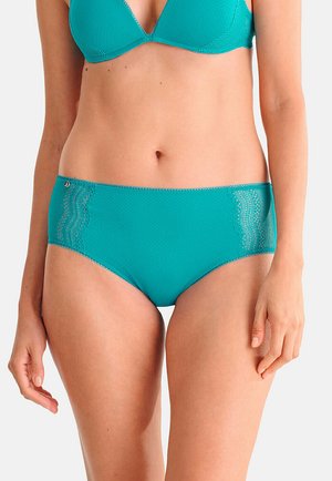 Femme portant un soutien-gorge vert sarcelle bordé de dentelle et une culotte taille haute assortie, debout les bras détendus le long du corps.