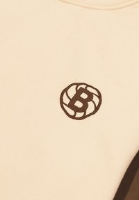 Sudadera beige con un logotipo bordado marrón que presenta un diseño circular y la letra "B" estilizada. Textura de tela suave, cuello redondo.