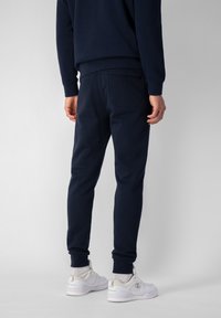 Pantalones de chándal azul marino con puños elásticos, cintura con cordón y un bolsillo trasero, combinados con zapatos deportivos blancos y calcetines claros.