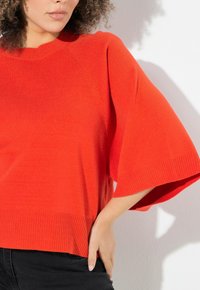 Roter gestrickter Pullover mit weiten Ärmeln und geripptem Saum. Die Stoffstruktur wirkt weich und leicht. Die Hand des Models ruht auf der Hüfte und zeigt die Passform.