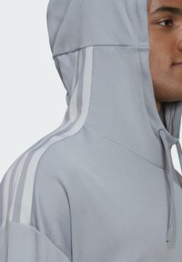 Lichtgrijze sportieve hoodie van een textuurstof, met drie witte verticale strepen langs de linkerschouder en een capuchon met koord.