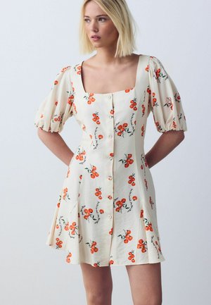 Woman wearing a cream button-down dress with puffed sleeves and orange cherry print, standing with hands behind her back against a plain background.