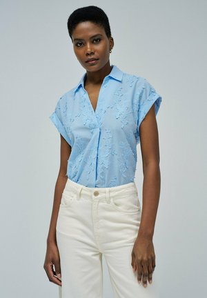 Blusa de manga corta azul claro con bordado floral, diseño con cuello y un corte relajado, combinada con pantalones de tiro alto en color blanco roto.