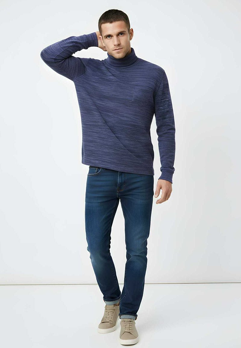 Jersey de cuello alto de punto en azul marino con un patrón texturizado, combinado con vaqueros azul oscuro y zapatillas deportivas beige claro. El modelo posa con una mano en la cabeza.