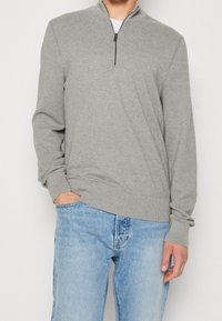 Man med en ljusgrå tröja med halvmatch och ribbade mudd och nederkant, kombinerat med ljusblå jeans, hand i fickan.