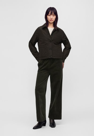 Femme debout les mains dans les poches, portant une veste boutonnée vert foncé, un pantalon large assorti et des bottines noires sur fond uni.