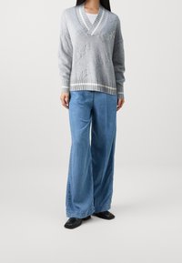 Hellgrauer gestrickter Pullover mit V-Ausschnitt und dezentem Muster, kombiniert mit weitem blauen Denim-Hosen und schwarzen quadratischen Schuhen.