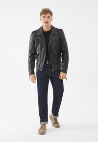 Giacca da motociclista in pelle nera con tasche con zip, dettagli testurizzati sulle spalle, indossata sopra una maglietta nera, abbinata a jeans scuri e scarpe beige.