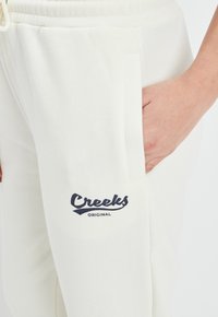 Beige Sweatpants aus weichem Material, mit seitlichen Taschen und einem navynblauen Logo-Druck auf dem Oberschenkel, der "Creeks ORIGINAL" liest.