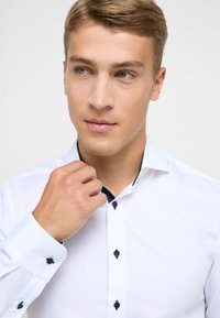 Witte overhemd met zwarte knopen en een gepatteerde marineblauwe kraag. Gladde textuur, aansluitend ontwerp en lange mouwen.