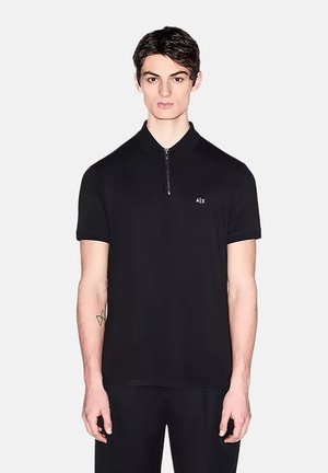 Jonge man met kort donker haar draagt een zwart poloshirt met korte mouwen, een rits aan de voorkant en een klein "Alx" logo op de borst.