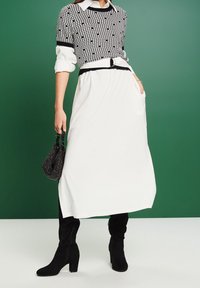 Top de punto negro y blanco con patrón, de manga larga, falda blanca, cinturón negro y botas negras hasta la rodilla, sosteniendo un pequeño bolso negro.