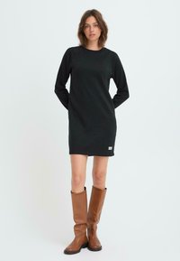 Musta college-mekko pitkillä hihoilla ja pyöreällä kaula-aukolla. Silkkinen pinta, polvipituinen, yhdistettynä ruskeisiin polvipitkiin nahkasaappaisiin.