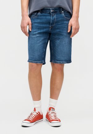 Άνδρας που φοράει μπλε τζιν σορτς μεσαίου μήκους, άσπρες κάλτσες και κόκκινα καμβά παπούτσια χαμηλού τύπου, στέκεται με τα χέρια χαλαρά δίπλα στο σώμα.