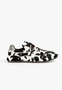 Zapatilla con estampado de vaca, textura de pelaje en blanco y negro, detalles en plata en el talón, cordones marrones y diseño de punta redondeada.