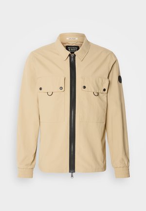 Beige jakke i let stof med en sort frontlynlås, to brystlommer og elastiske manchetter. Har mærketikette indeni kraven.