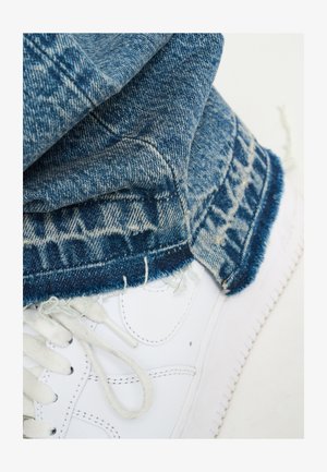 Bílé sneakersy s texturovanými tkaničkami, spárované s modrými džínami s roztrhaným lemem a geometrickým vzorem blízko manžety.