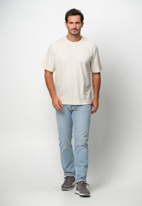 Beżowy t-shirt z krótkim rękawem i okrągłym dekoltem, w połączeniu z jasnoniebieskimi dżinsami i szarymi sneakersami. Prosty, jednolity design bez wzorów.