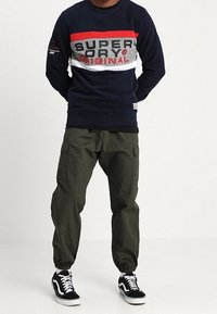 Uomo che indossa una felpa blu marina con strisce rosse, grigie e bianche e testo, pantaloni cargo oliva e sneakers nere e bianche, in piedi con le mani dietro la schiena.