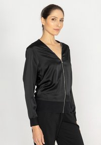 Czarna satynowa kurtka bomberka z głębokim dekoltem w kształcie V, zamkiem z przodu i ribbowymi mankietami. Gładka faktura i luźny krój zwiększają komfort i styl.