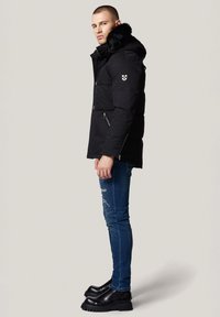Czarna kurtka puchowa z kapturem wyłożonym futrem, kieszeniami na zamek i detalem z logo. Nosi się ją z przetartymi niebieskimi dżinsami i czarnymi butami na platformie.