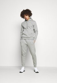 Grau Baumwollhoodie mit Kordelzug, kombiniert mit passenden tapered Jogginghosen. Beide zeigen dezente Logoakzente. Tragen Sie dazu weiße Sportschuhe.