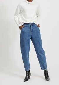 Niebieskie, jeansowe spodnie z wysokim stanem o luźnym kroju, z plisami z przodu i krótką długością, połączone z białym sweterkiem z prążkowanej dzianiny i czarnymi botkami za kostkę.