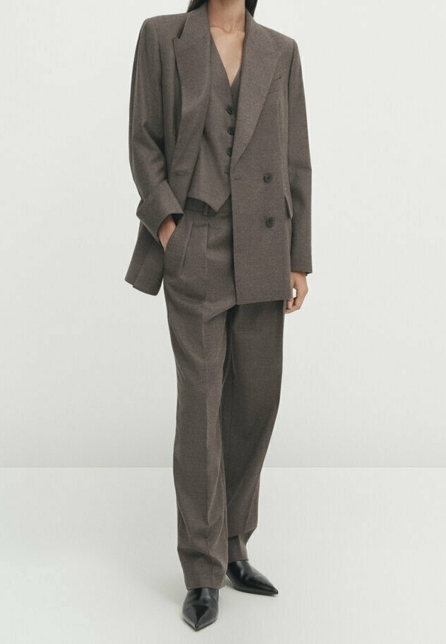 Femme portant un costume trois pièces marron ajusté avec un pantalon à jambes larges et des chaussures noires à bout pointu, debout devant un fond clair uni.