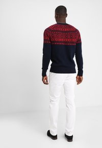 Marineblauwe trui met een rood geometrisch patroon over de schouders, ribgebreide mouwen en zoom. Gepaarde met witte broek en zwarte schoenen.