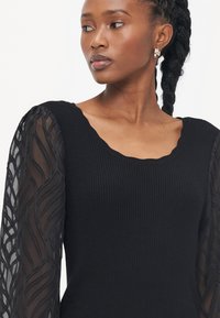 Zwarte geribbelde trui met een scalloped halslijn en sheer, textuur-handen met een golvend patroon. Een fitted silhoeët accentueert de vorm.