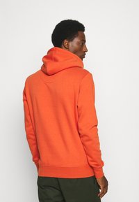 Mężczyzna w jaskrawo pomarańczowej bluzie z kapturem i ciemnozielonych spodniach, stojący i patrzący na bok na tle jednolitego jasnego tła.