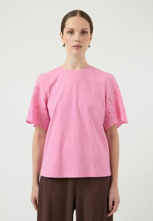 Naine, kes kannab roosat pluusi lühikeste pitsiliste varrukatega ja pruune pükse, seisab ühevärvilise heleda tausta ees.