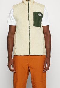 Uomo che indossa un gilet in pile color crema con tasca sul petto verde scuro, camicia bianca a maniche corte e pantaloni arancioni, in piedi contro uno sfondo bianco.