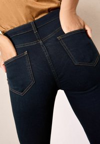 Vaqueros azul oscuro de cintura alta, con bolsillos de parche, costuras amarillas en contraste y una textura suave.