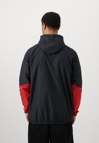 Mand med kort, krøllet hår iført en sort hættetrøje med røde ærmer, set fra baggrunden mod en lys, ensfarvet baggrund.